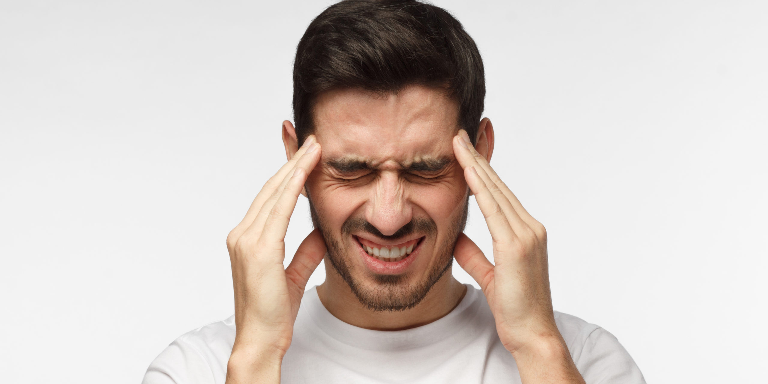 Portrait of young man isolated on grey background suffering from severe headache, pressing fingers to temples, closing eyes to relieve pain with helpless face expression