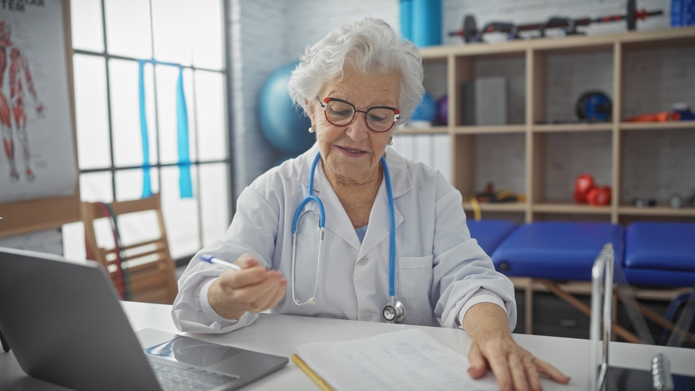 Senior,Woman,Doctor,Writing,In,A,Clinic,Office,With,Laptop,
