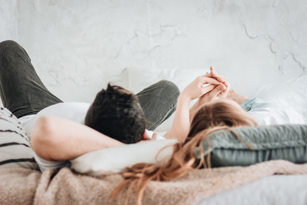 Young,Straight,Couple,Laying,On,Backs,On,Bed,Holding,Hands.
